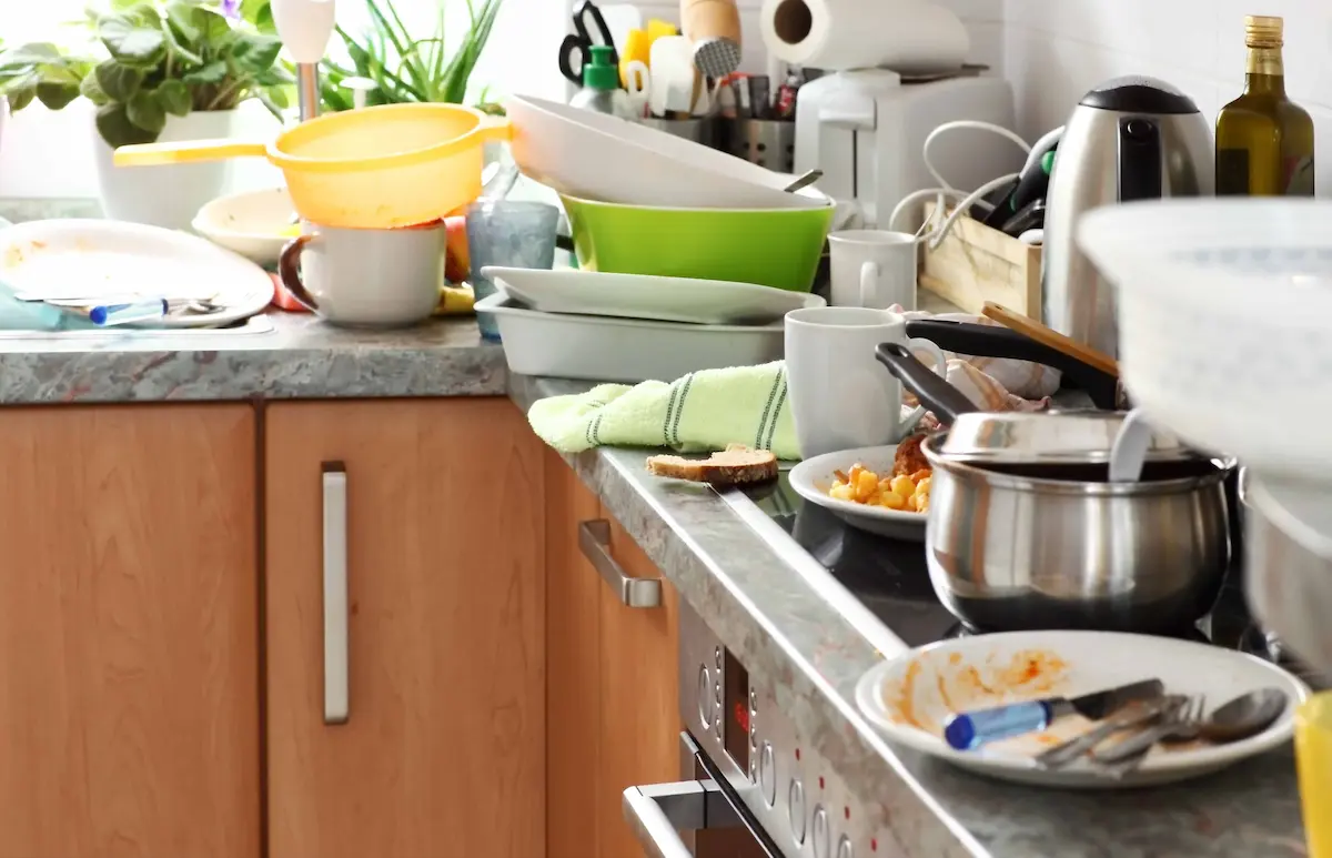 Messy kitchen countertop with dirty dishes, cookware, cups, and food leftovers waiting to be cleaned