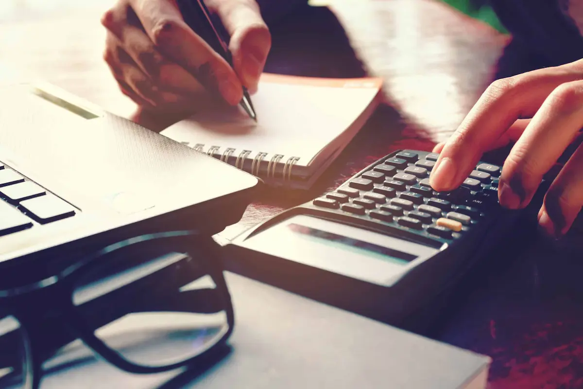 Person using calculator and writing financial notes beside laptop
