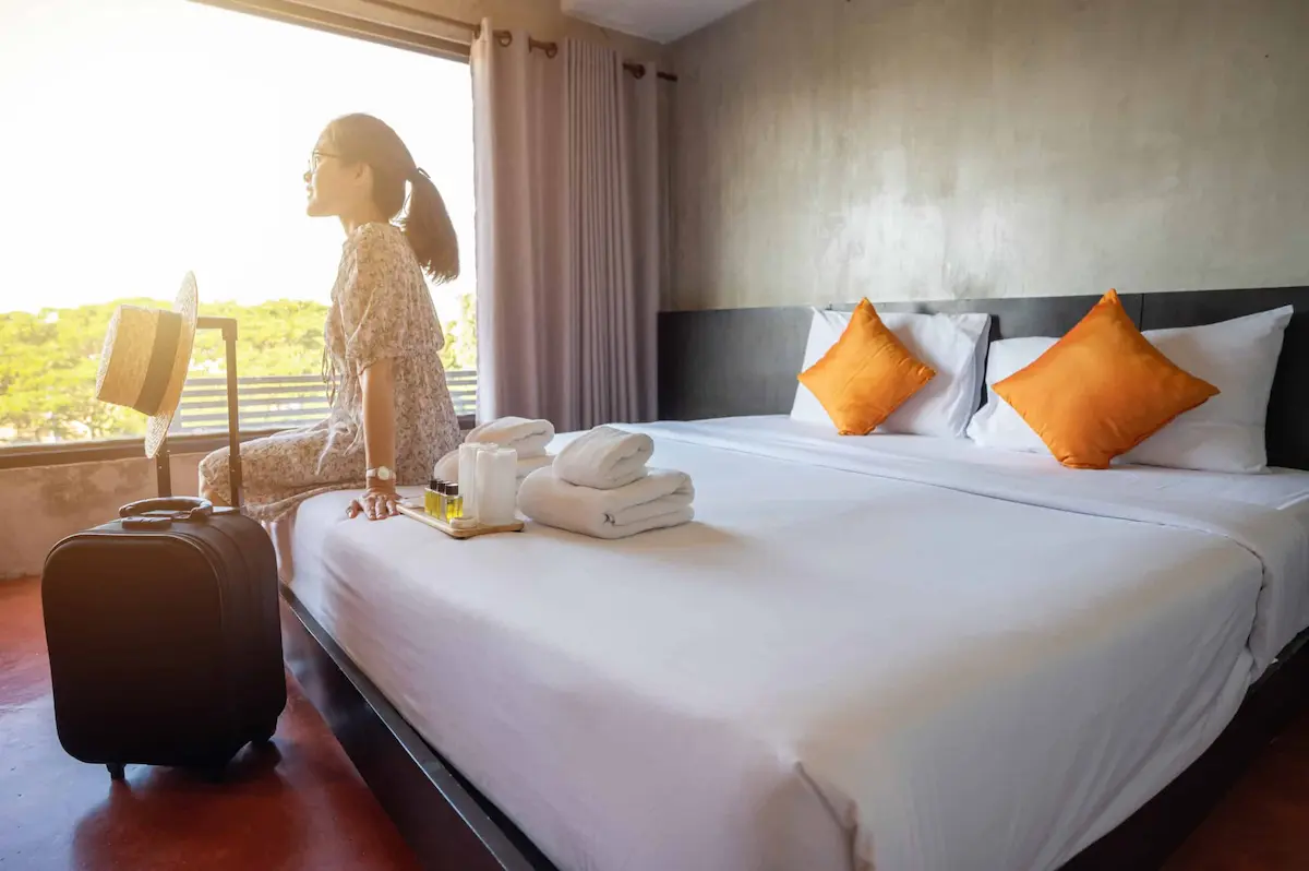 Traveler sitting on hotel bed beside luggage near bright window