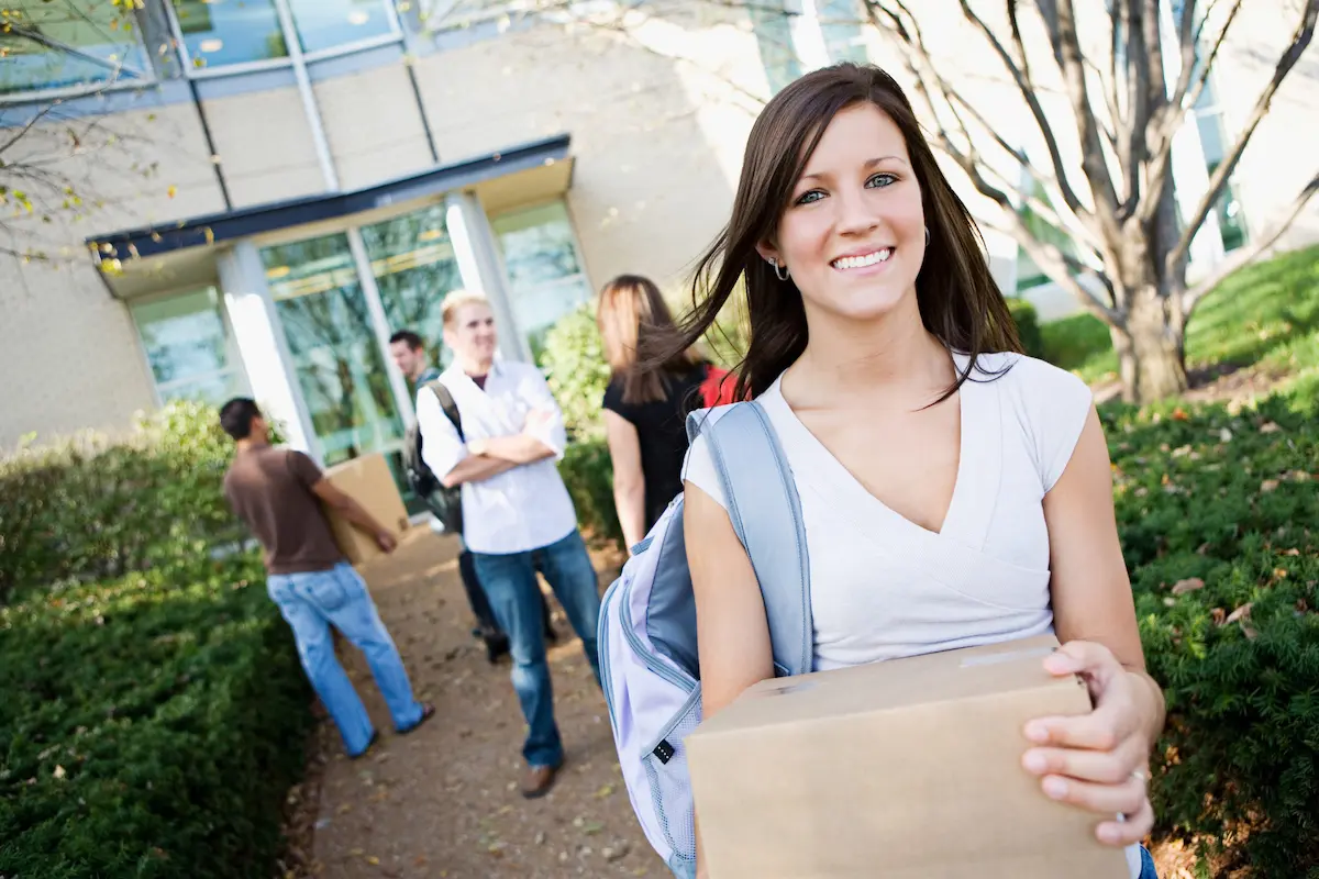 Smiling college student carrying a moving box outside campus housing